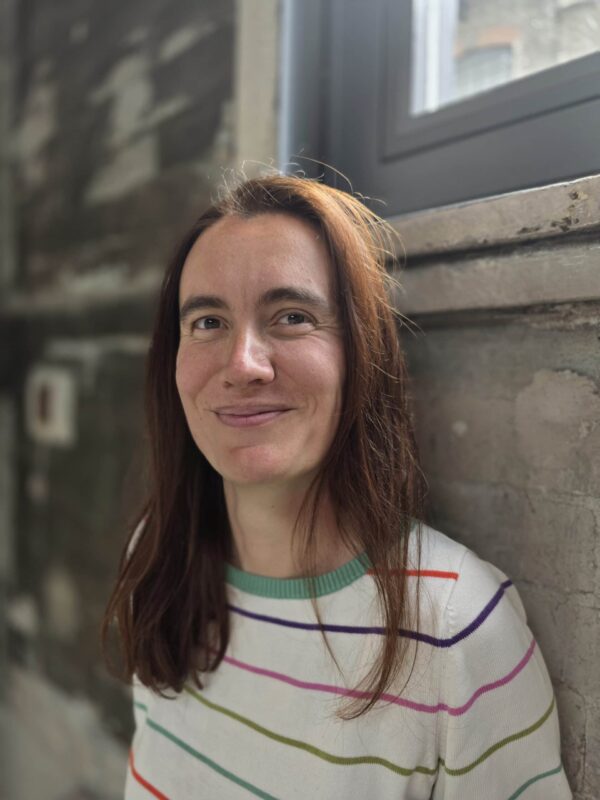 Frau mit braunen Haaren vor einer Steinmauer und Fenster.